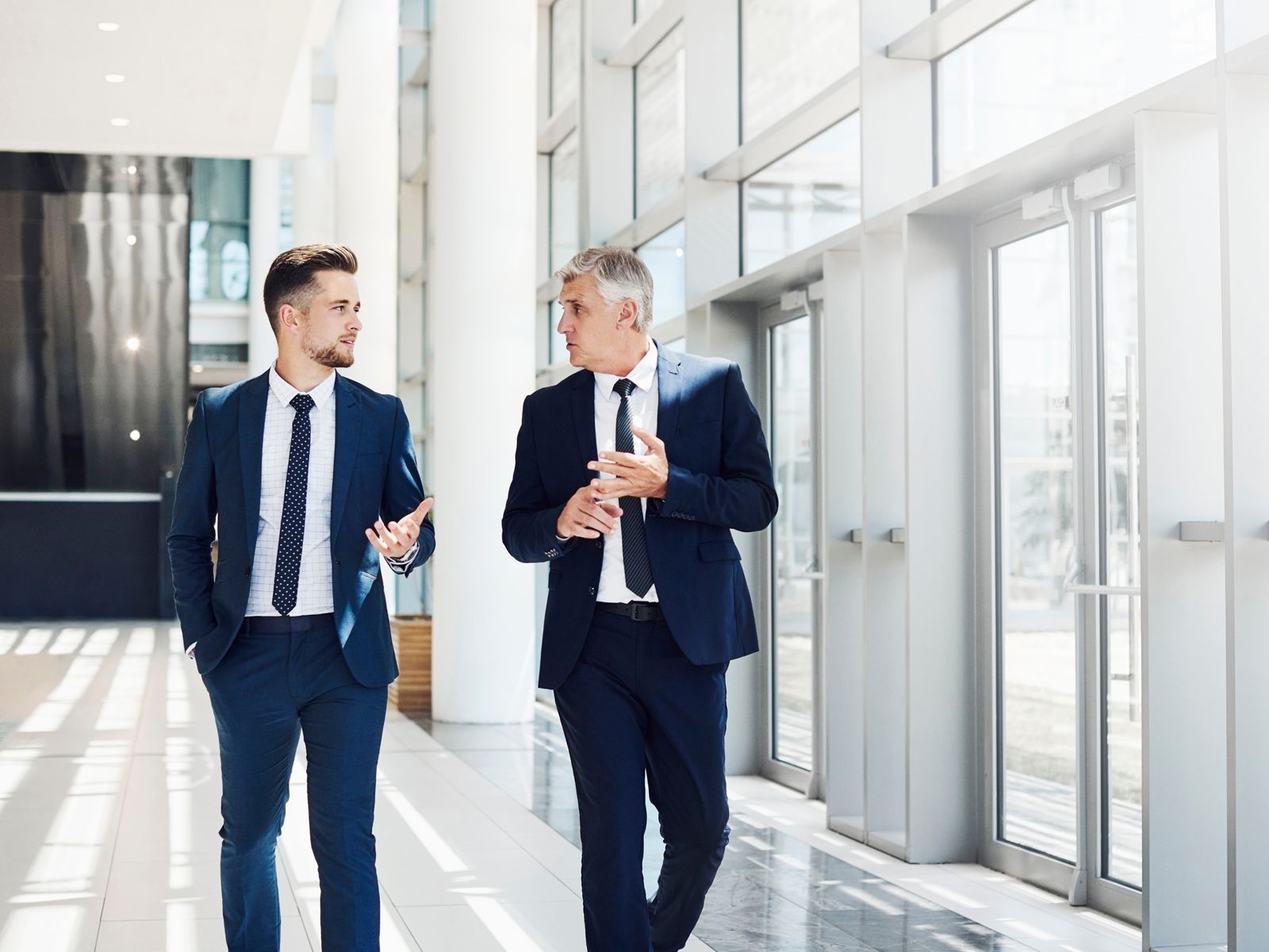 Two men in suits and ties are walking down a hallway and talking.